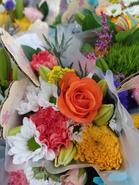 A vibrant floral bouquet featuring an orange rose at the center, surrounded by pink carnations, white daisies with green centers, yellow chrysanthemums, and small purple lavender sprigs. The arrangeme