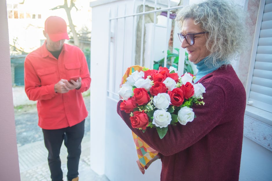 A woman with curly gray hair, wearing glasses, a blue turtleneck, and a burgundy sweater, holding a vibrant floral bouquet featuring red and white roses with green foliage, wrapped in decorative patte