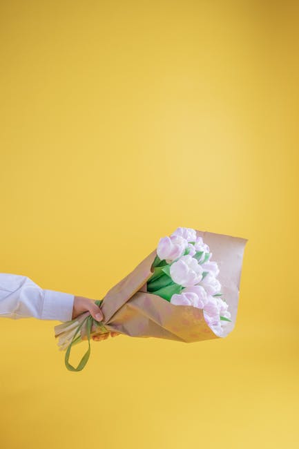 A person wearing a white shirt is holding a bouquet of soft pink tulips wrapped in light brown paper and tied with a green ribbon, offering a fresh floral gift. The tulips have smooth, rounded petals