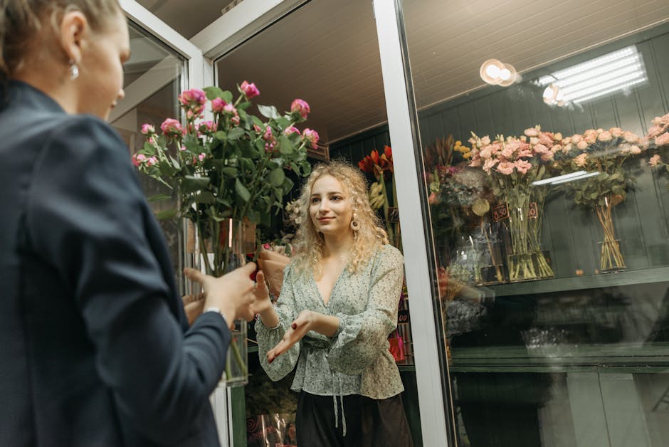 A young woman with curly blonde hair and light makeup receives a bouquet of blooming pink roses wrapped in clear cellophane from a florist at a flower shop near Mill Hill Broadway station. The florist