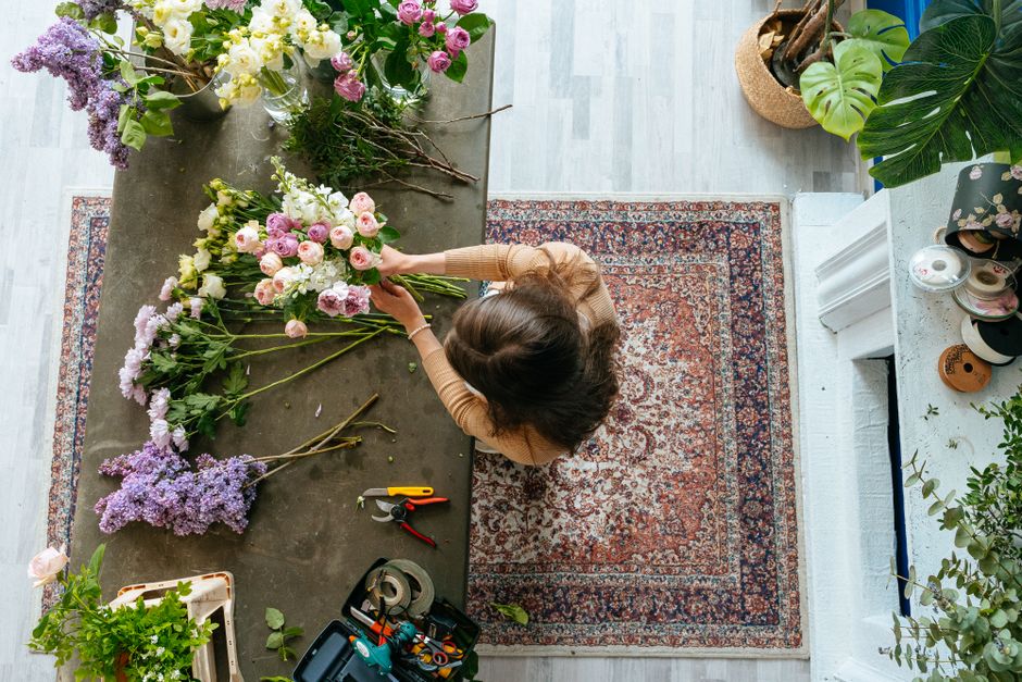 A florist with shoulder-length brown hair arranging a bouquet of fresh flowers on a dark grey work table inside a well-lit room. The bouquet includes pink, white, and purple flowers, with some flowers