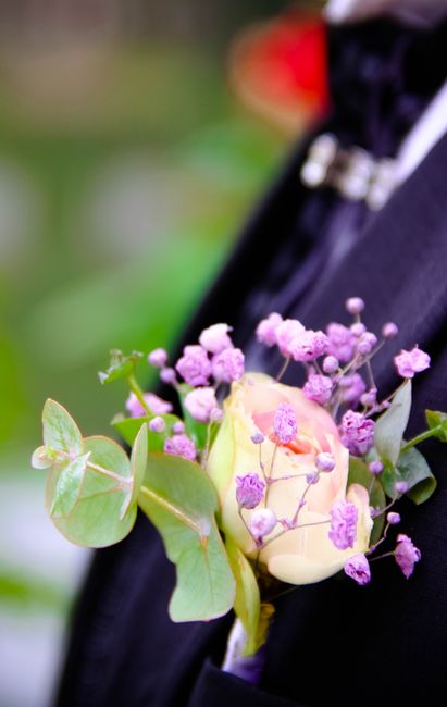 A floral boutonniere featuring a single pale pink rose surrounded by small purple gypsophila flowers and green eucalyptus leaves, arranged in a compact cluster and attached to a dark suit lapel with a