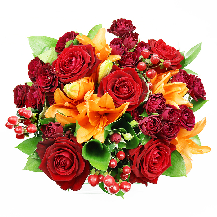 Viewed from a top-down perspective against a pristine white surface, this opulent bouquet is arranged in a dense, perfect round that radiates warmth and celebration. The eye is drawn to large, fully blossomed deep red roses whose velvety petals curl and overlap to create sensual, layered centres suggestive of classic romance. Interspersed amid them are vivid orange Asiatic lilies, their trumpet-shaped petals and prominent stamens offering bold architectural moments; some lilies are fully open, others in partial unfurling or tight bud form, adding a sense of unfolding life. Nestled between the dominant flowers are clusters of darker burgundy spray roses, miniature in scale but rich in texture, creating shadowed pockets that deepen the palette. Bright red hypericum berries, smooth and glossy, punctuate the composition with jewel-like points that reflect the soft studio lighting. Tucking everything together are broad, glossy ruscus or salal leaves whose cool green tones provide contrast and a natural, structural framework. The gentle, even lighting highlights surfaces-the satin sheen of petals, the tactile grain of rose edges, the glassy gloss of berries-without harsh contrast, helping to communicate colour and form to someone relying on description. The arrangement smells faintly of lilies' spicy citrus and the warm, honeyed scent of freshly cut roses. I imagine this bouquet on a windowsill near Hale Lane or delivered to a reception at a Mill Hill venue; it suits anniversaries, autumnal celebrations, or an elegant gesture meant to convey affection and joy.